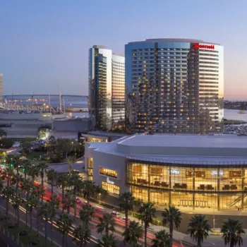 A large hotel complex with glass towers and a brightly lit building in front, adjacent to a marina, at dusk.