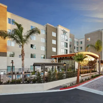 A modern, multi-story hotel with palm trees and landscaped grounds under a clear sky at sunset; entrance features a covered driveway.