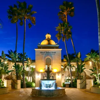 A hotel building with a central dome and bell tower is illuminated at night, surrounded by palm trees and a fountain in the foreground.