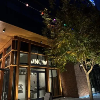 A modern storefront with a sign reading "THE NOW" is lit at night, with a sandwich board outside and a tree decorated with colorful lights nearby.