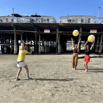 Three women pose with beach balls under a wooden pier on a sandy beach; one takes a photo while the other two stand with their arms raised.