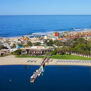Aerial view of a beachfront resort with a sandy beach, dock, lounge chairs, palm trees, and buildings, with the ocean and cityscape in the background.