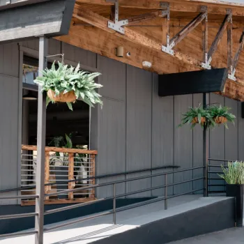 Modern building exterior with exposed wooden beams, hanging plants, a ramp for accessibility, and a planter with tall greenery near the entrance.
