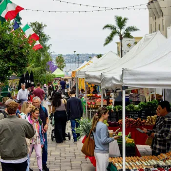 People walk through an outdoor market with white tents, fresh produce on display, and various flags hanging overhead on a cloudy day.