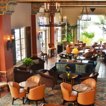 Elegant hotel lounge with armchairs and sofas, marble tables, chandeliers, large windows, potted plants, and a grand piano, featuring colorful and ornate decor.