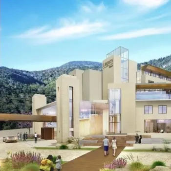 Modern multi-story hotel building with glass accents, set against a forested hillside; people walk on the path and cars are parked near the entrance.