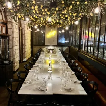 Long dining table set for a meal in a restaurant with black chairs, marble tabletops, and a ceiling decorated with hanging greenery and lemons. Warm lighting creates a cozy atmosphere.