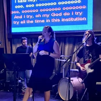 A band performs on stage with a woman singing at the center. Musicians play guitar and drums. Karaoke lyrics are displayed on a large screen in the background.