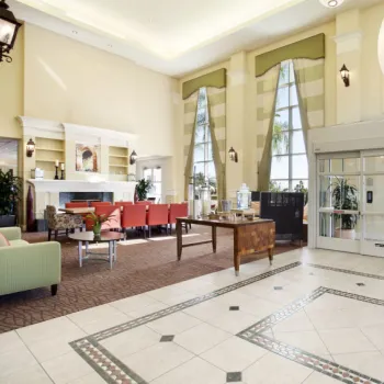 Spacious hotel lobby with tiled floors, large windows, a seating area with red chairs, a fireplace, and double glass entrance doors.