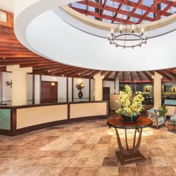 Spacious hotel lobby with tiled floor, round skylight, central table displaying floral arrangement, seating area, and reception desk in the background.