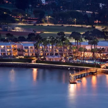 A waterfront hotel with a pool is illuminated at dusk, surrounded by palm trees and facing a dock that extends into calm water. Sports fields and buildings are visible in the background.