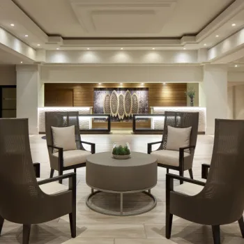 Modern hotel lobby with four high-back chairs arranged around a circular table, neutral tones, and a reception desk in the background.
