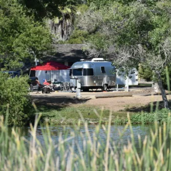 A silver travel trailer is parked at a campsite with a red canopy and chairs nearby, surrounded by trees and greenery by the edge of a pond.