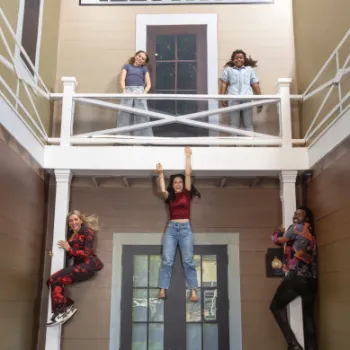 Four people pose on an illusion set at the Museum of Illusions, with two appearing to hang off a balcony and two standing above, creating a visual trick.