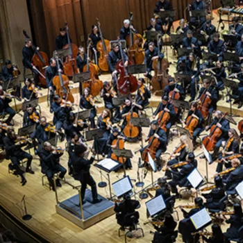 An orchestra performs on stage, with musicians playing various instruments and a conductor leading at the front.