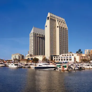 Tall modern hotel buildings overlook a marina with several docked boats under a clear blue sky.