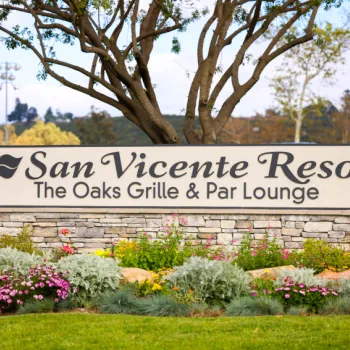 A sign in front of landscaping reads "San Vicente Resort The Oaks Grille & Par Lounge" with trees and stone wall behind it.
