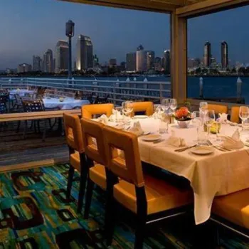 A dining table set for eight is positioned by large windows overlooking a city skyline and waterfront at dusk.