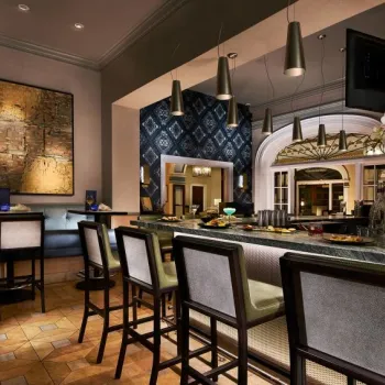 Modern hotel bar with high-backed chairs, a marble counter, pendant lighting, patterned wallpaper, and various drinks and snacks arranged on the counter.
