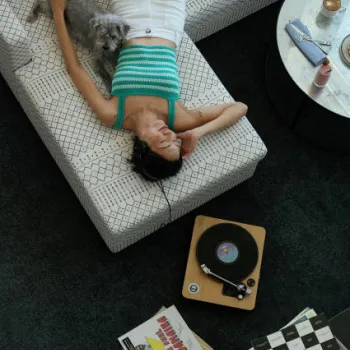 A woman in a blue striped top lies on a sofa with a dog, listening to music on headphones; a record player and vinyl records are on the table nearby.