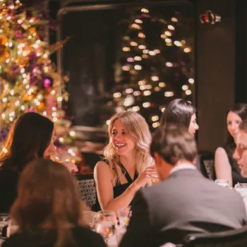 A group of people sit at a dinner table in a dimly lit room with a decorated Christmas tree and string lights in the background.