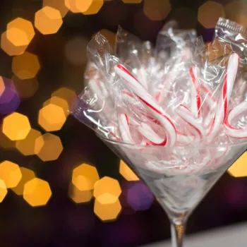 A glass martini glass holds multiple individually wrapped mini candy canes, with out-of-focus festive lights in the background.