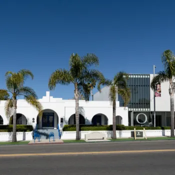 A modern white building with large arched windows and palm trees in front, set against a clear blue sky.