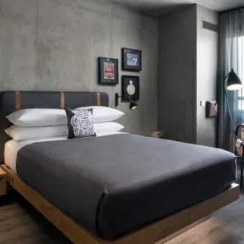 Modern hotel room with a grey concrete wall, double bed with dark bedding, wooden side table, wall art, armchair by the window, and floor lamp. Natural light filters through sheer curtains.