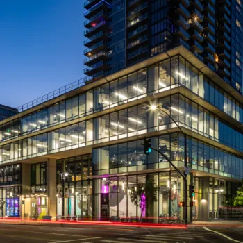 Modern glass office building and high-rise at dusk, with illuminated interiors and light trails from passing vehicles on city streets.