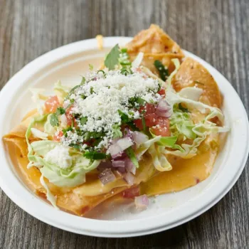 A plate of nachos topped with melted cheese, lettuce, diced tomatoes, onions, cilantro, and crumbled cheese on a wooden table.