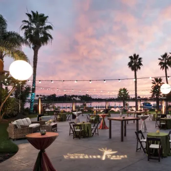 Outdoor event space at sunset with string lights, palm trees, round tables with green linens, and a waterfront view in the background.