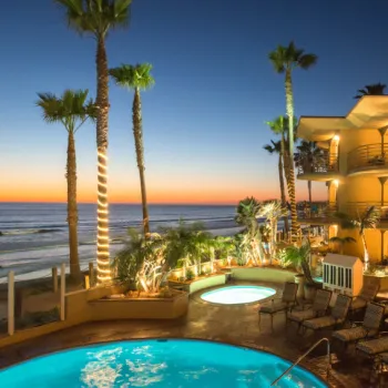 Hotel pool area with lounge chairs, hot tub, and palm trees, overlooking the ocean at sunset with building lights illuminated.