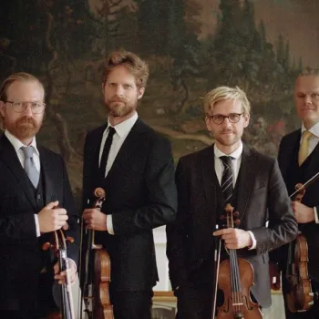 Four men in suits stand indoors holding string instruments, including violins and a viola, in front of a large, classical-style painting.
