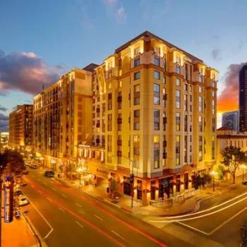 A brightly lit urban building stands at a street corner at dusk as cars create light trails; high-rise buildings and a colorful sunset sky are visible in the background.