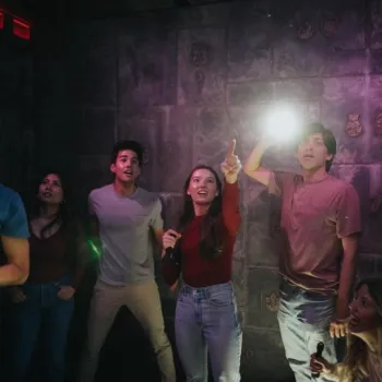 A group of people in a dimly lit room look around with flashlights, appearing to search or solve a puzzle on stone-embellished walls.