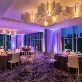 A modern event space with round tables set for dinner, white chairs, large windows, and geometric ceiling lights, illuminated by purple and warm lighting.