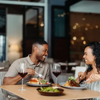 Two people sit at a restaurant table, eating dinner and drinking red wine while smiling at each other.