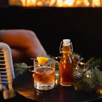 A glass of cocktail with a lemon twist and a small bottle on a table decorated with pine branches and lights; blurred fireplace and seated person in the background.