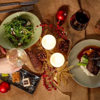 Overhead view of a holiday dinner with salad, steak, wine, and desserts on a brown tablecloth, decorated with candles, pinecones, and ornaments. Two people are dining.