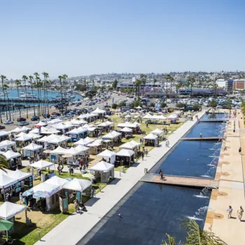 A waterfront outdoor market with white tents, palm trees, walkways, and reflecting pools, with a marina, roads, and city buildings in the background under a clear sky.