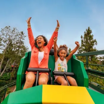 Two people, an adult and a child, are riding a green and yellow roller coaster outdoors, raising their hands and smiling. Trees and a blue sky are in the background.