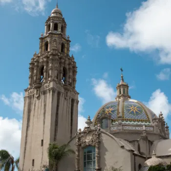 A historic building with an ornate bell tower and a dome decorated with colorful tiles, set against a blue sky with scattered clouds.