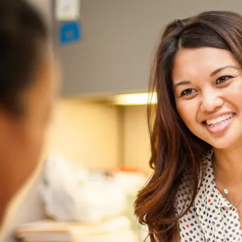 A woman sits at a desk, smiling and talking to another person in an office setting.