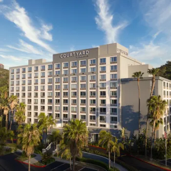 Exterior view of a multi-story Courtyard by Marriott hotel with palm trees in front, set against a backdrop of hills and a partly cloudy sky.