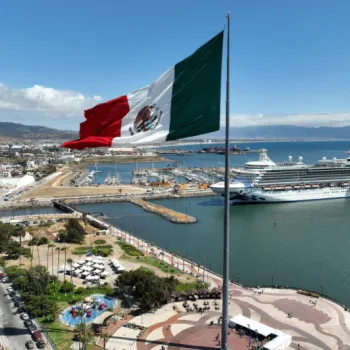 A large Mexican flag stands by the waterfront in a city port with a cruise ship docked nearby and urban buildings visible in the background.