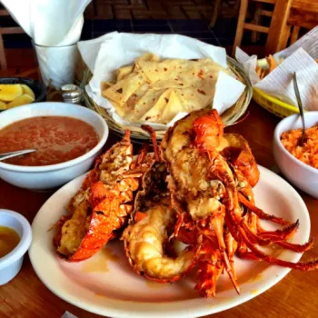 A plate of cooked lobster with melted butter, served with rice, beans, tortillas, lemon wedges, and chips on a wooden table.