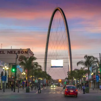 A large arch stands at the center of a city street at sunset, with cars, palm trees, shops, and Hotel Nelson visible on either side.