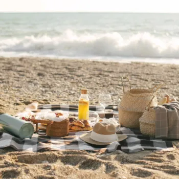A picnic setup with food, drinks, hats, and baskets on a blanket on a sandy beach near the ocean waves.
