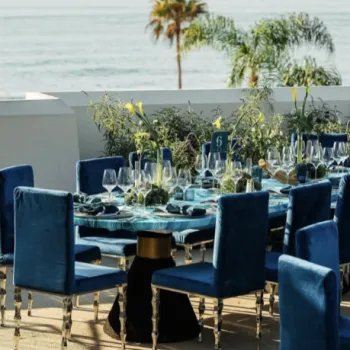 A formal dining table set with blue velvet chairs, glassware, and greenery centerpieces on an outdoor terrace overlooking the ocean.