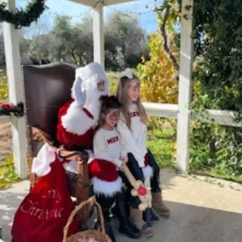 Santa Claus sits in a chair with two young girls beside him, all dressed in festive attire. A basket of candy canes is on the ground in front. The setting is outdoors under a canopy.